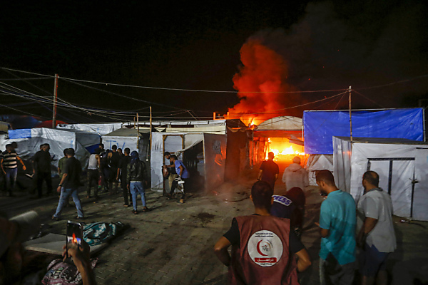 Auf dem Bild sieht man Menschen und Zelte in der Nacht in einem palästinensischen Flüchtlings-Lager im Gaza-Streifen. Im Hintergrund sieht man ein Feuer nach einem israelischen Luftangriff.