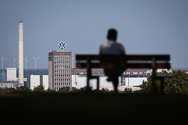 Auf dem Bild sieht man einen Mann auf einer Bank sitzen. Im Hintergrund sieht man Gebäude des VW-Hauptsitzes in Wolfsburg mit dem VW-Logo.