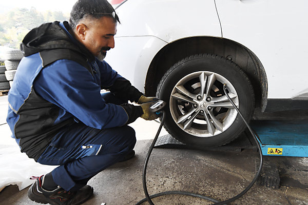 Auf dem Bild sieht man einen Mechaniker in einer Werkstatt. Er misst gerade den Druck bei einem Auto-Reifen.