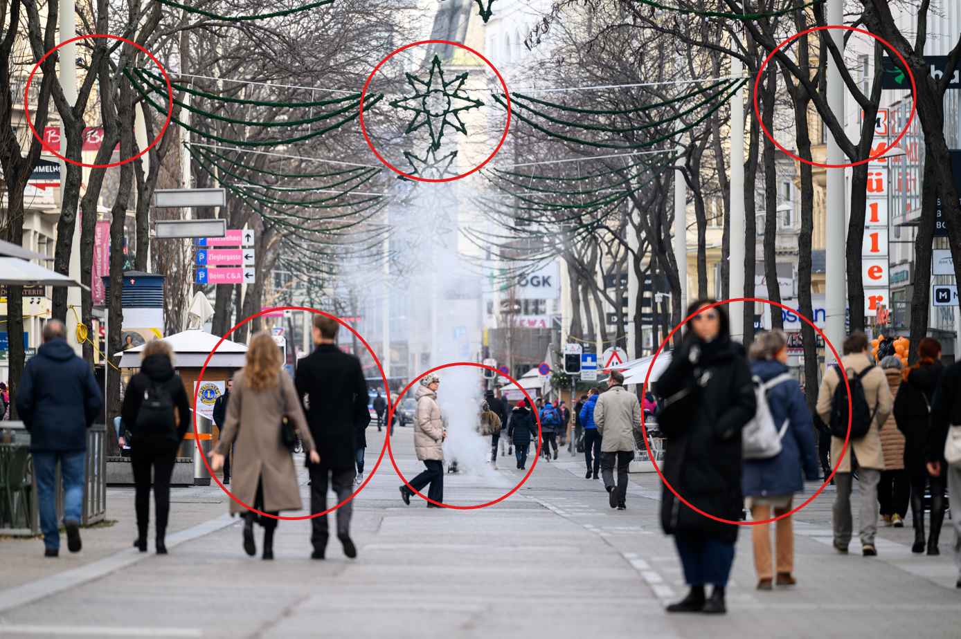 Mehrere Passanten mit warmer Kleidung auf der Mariahilfer Straße. Im Hintergrund dampft es aus einem Kanaldeckel. Man sieht kahle Bäume und Weihnachtsbeleuchtung.